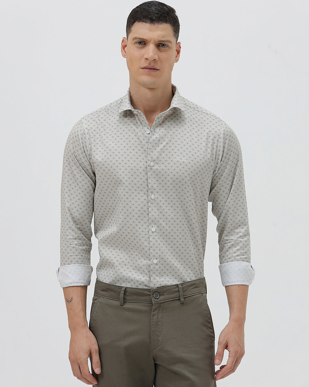 A man wearing a light gray long-sleeve button-down shirt with a small repeating geometric pattern, paired with olive green trousers, against a plain light gray background.