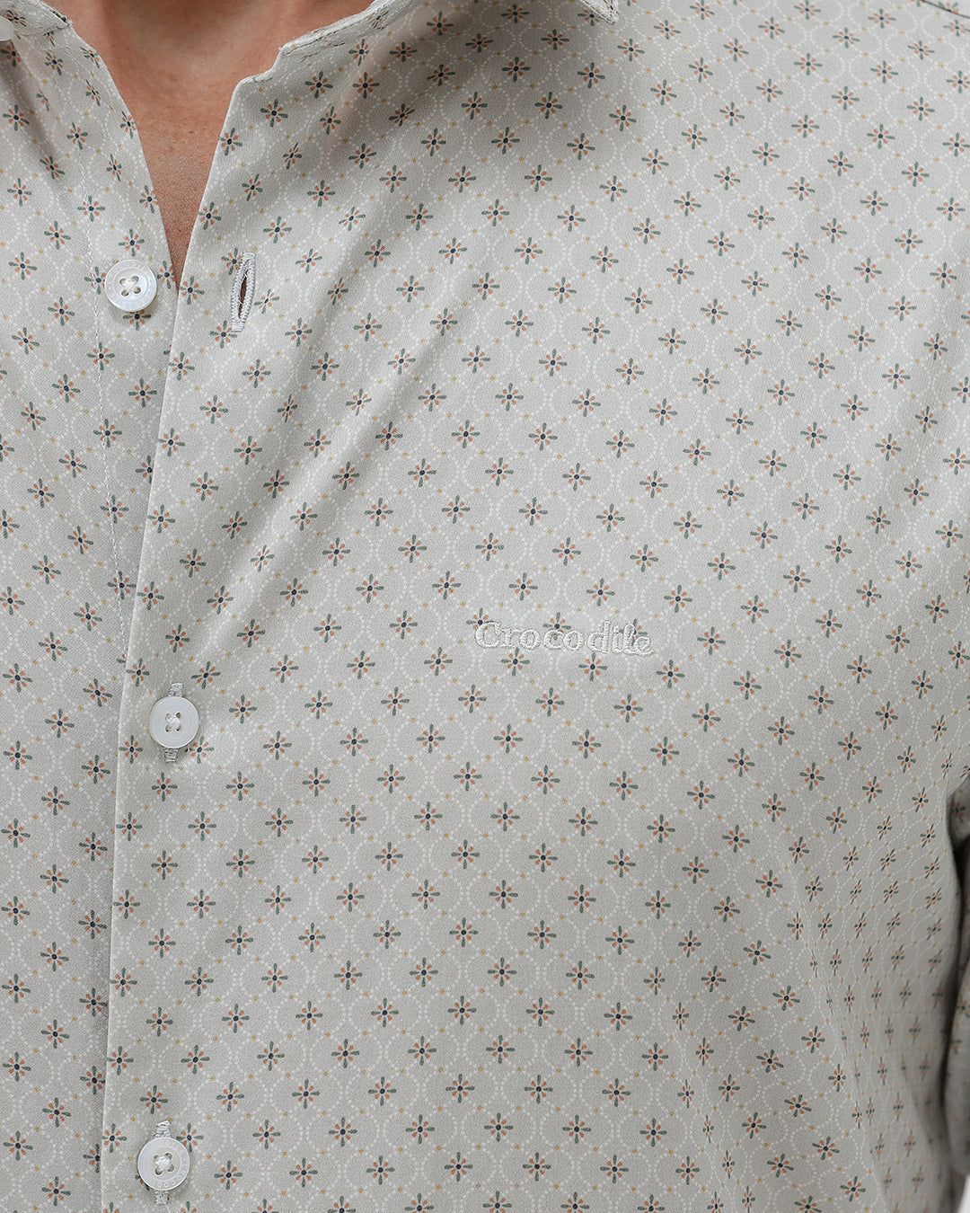 A man wearing a light gray long-sleeve button-down shirt with a small repeating geometric pattern, paired with olive green trousers, against a plain light gray background.