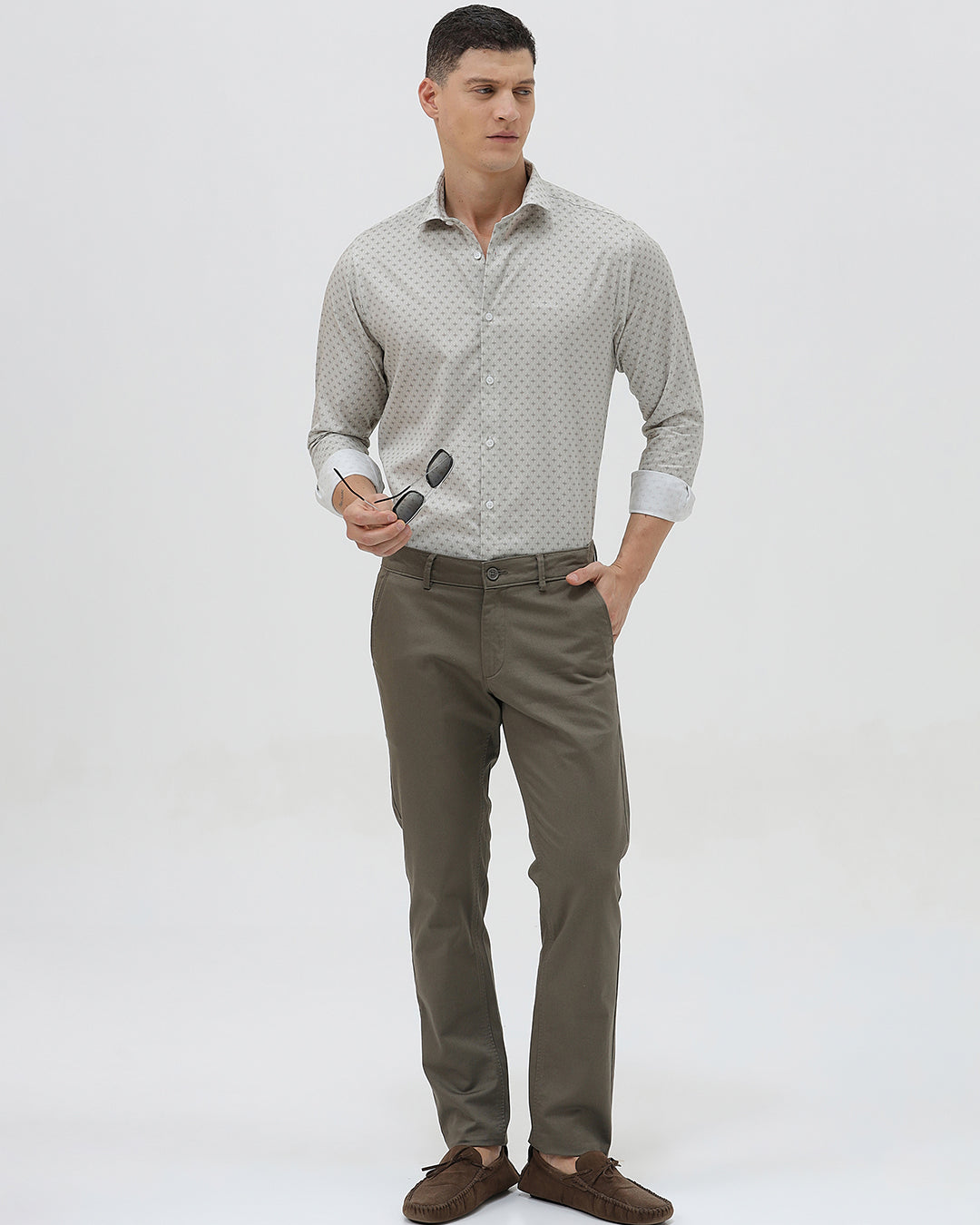 A man wearing a light gray long-sleeve button-down shirt with a small repeating geometric pattern, paired with olive green trousers, against a plain light gray background.
