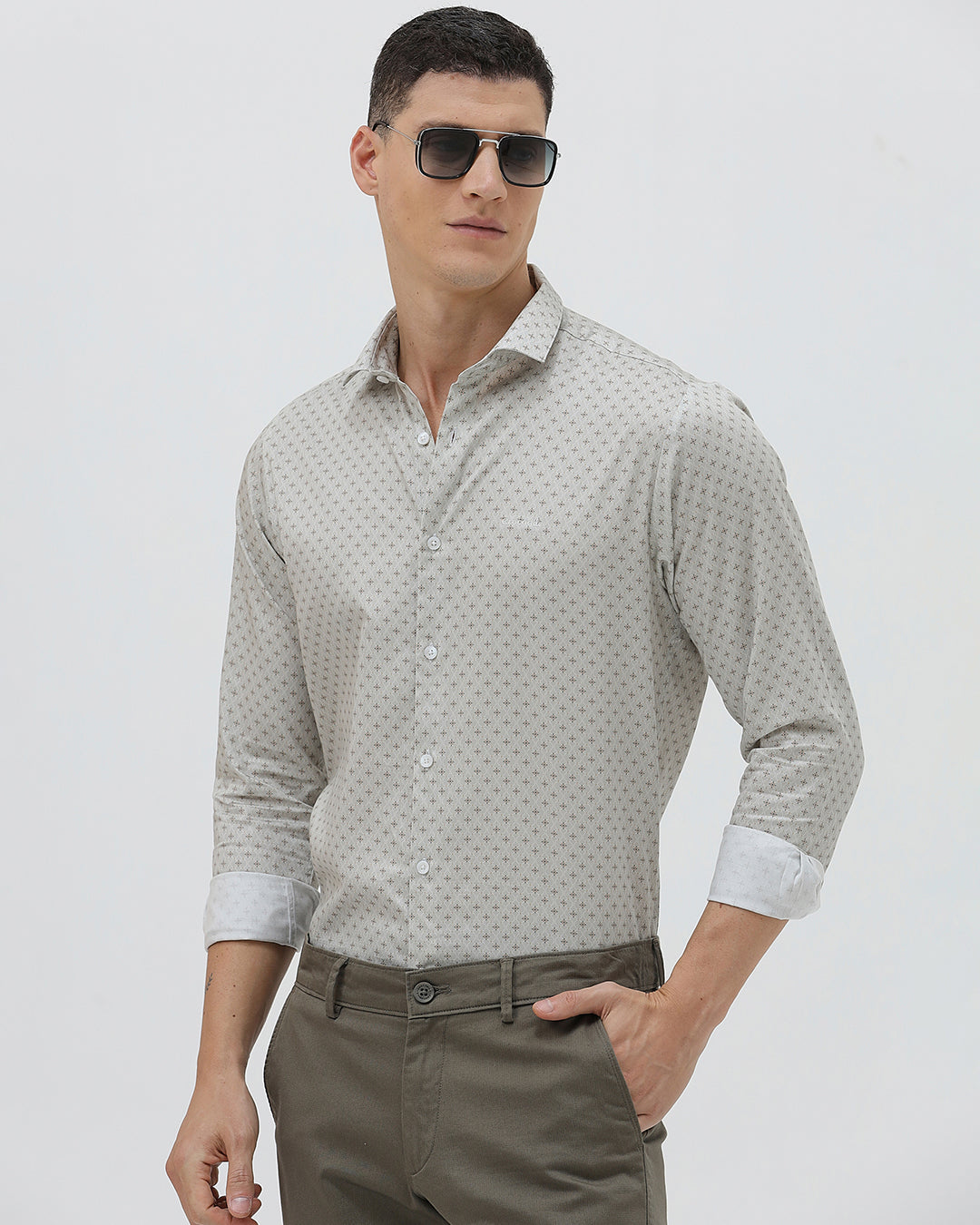 A man wearing a light gray long-sleeve button-down shirt with a small repeating geometric pattern, paired with olive green trousers, against a plain light gray background.