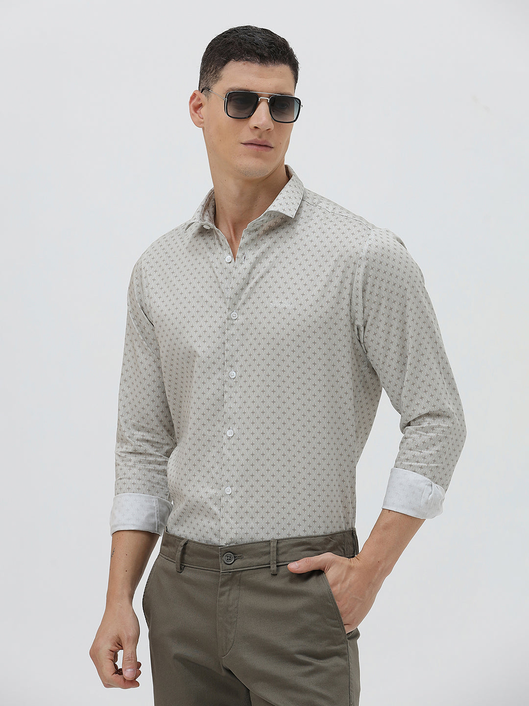 A man wearing a light gray long-sleeve button-down shirt with a small repeating geometric pattern, paired with olive green trousers, against a plain light gray background.