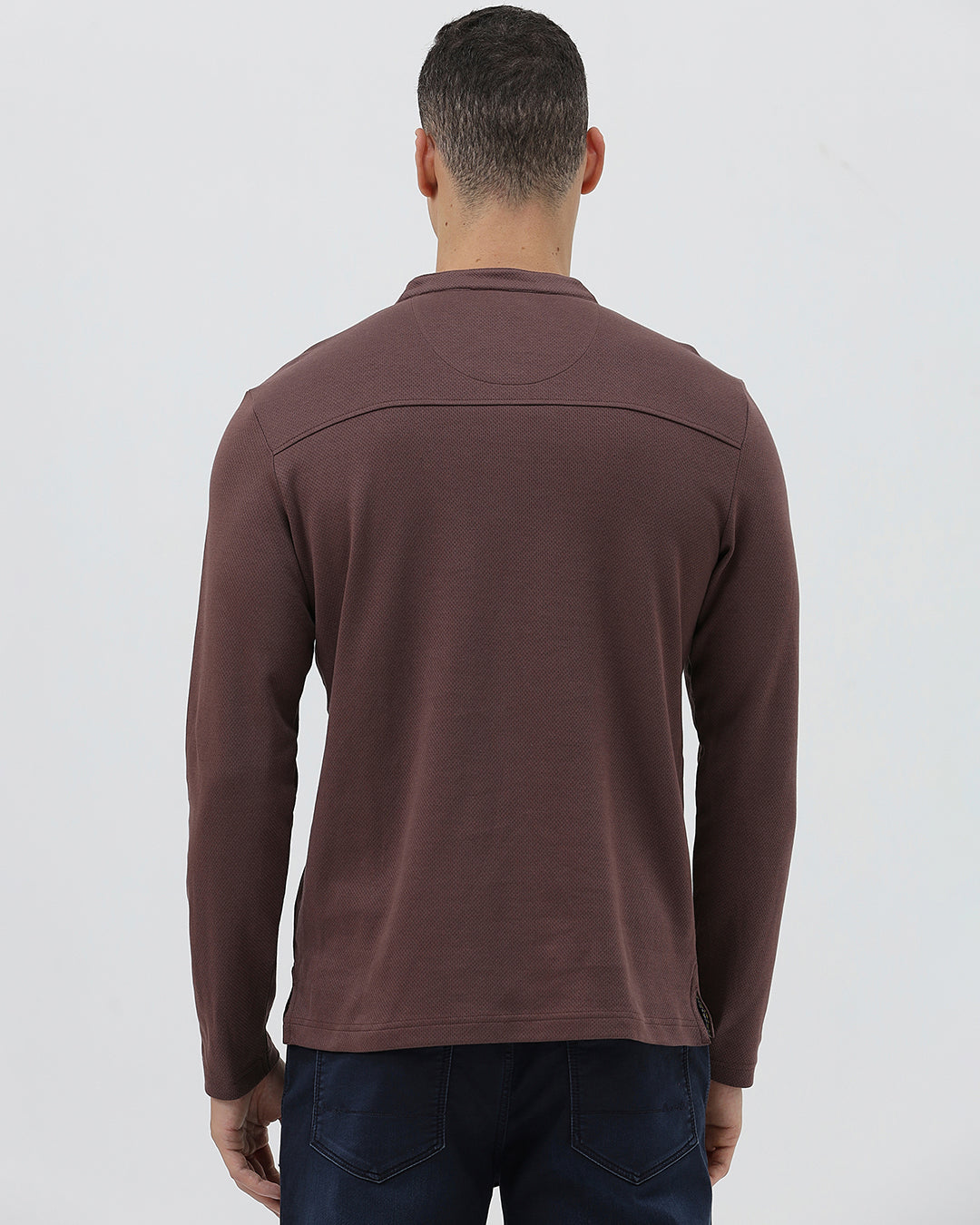 A man wearing a brown long-sleeve henley shirt with a small embroidered logo on the left chest, paired with dark blue jeans, standing against a light gray background.