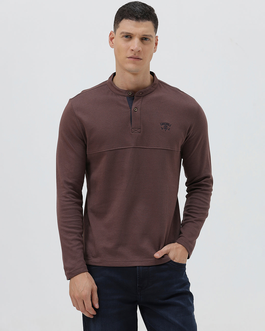 A man wearing a brown long-sleeve henley shirt with a small embroidered logo on the left chest, paired with dark blue jeans, standing against a light gray background.