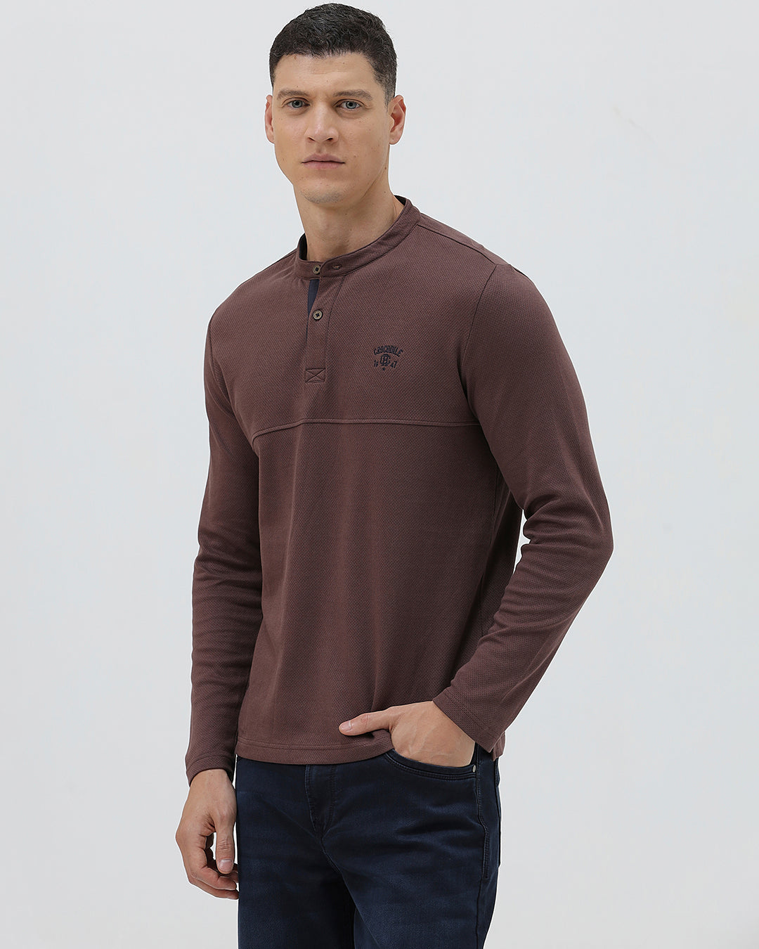 A man wearing a brown long-sleeve henley shirt with a small embroidered logo on the left chest, paired with dark blue jeans, standing against a light gray background.