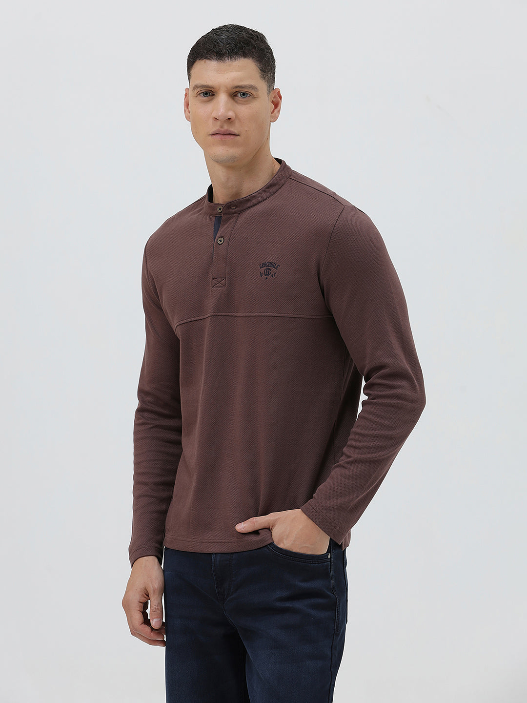 A man wearing a brown long-sleeve henley shirt with a small embroidered logo on the left chest, paired with dark blue jeans, standing against a light gray background.