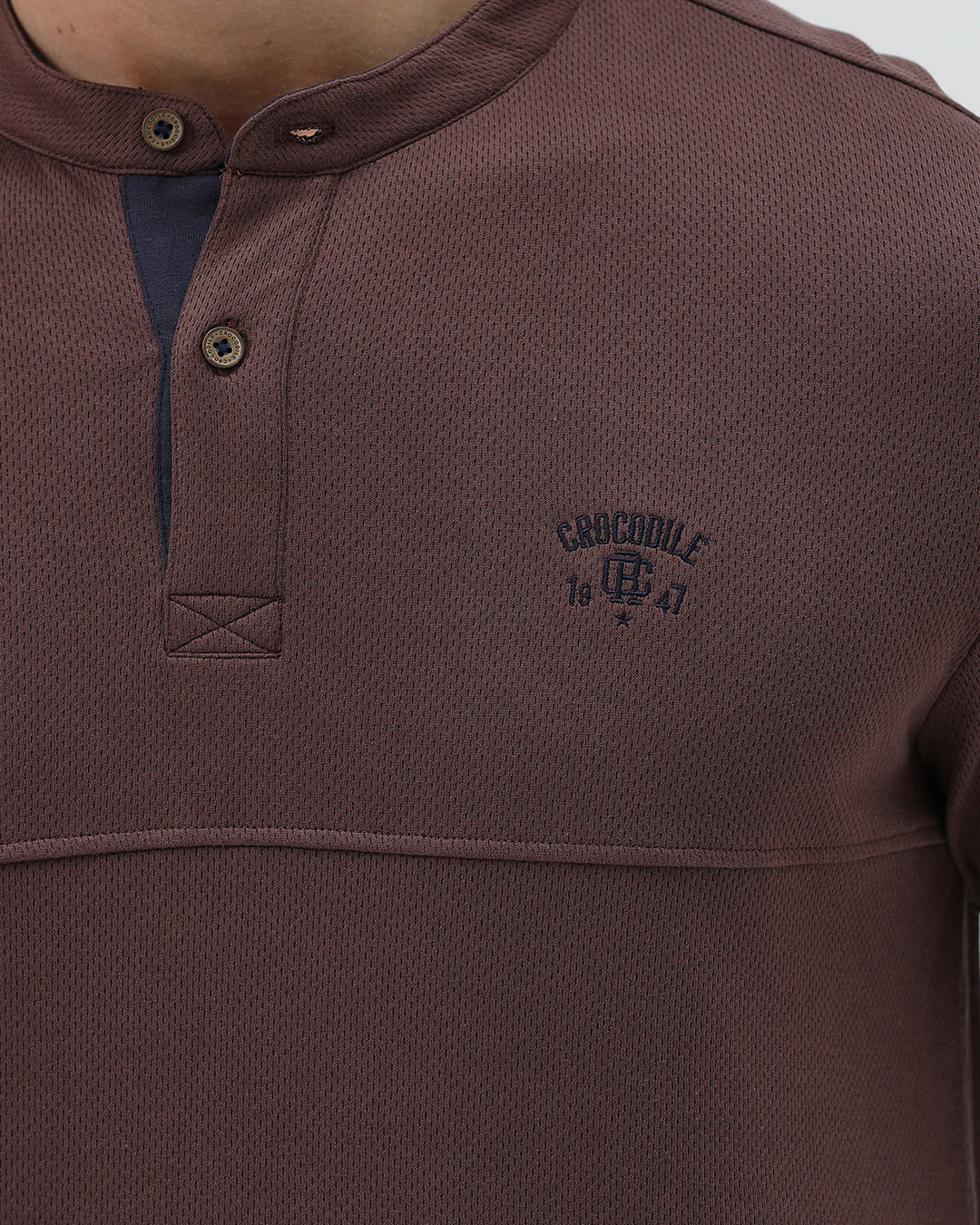 A man wearing a brown long-sleeve henley shirt with a small embroidered logo on the left chest, paired with dark blue jeans, standing against a light gray background.