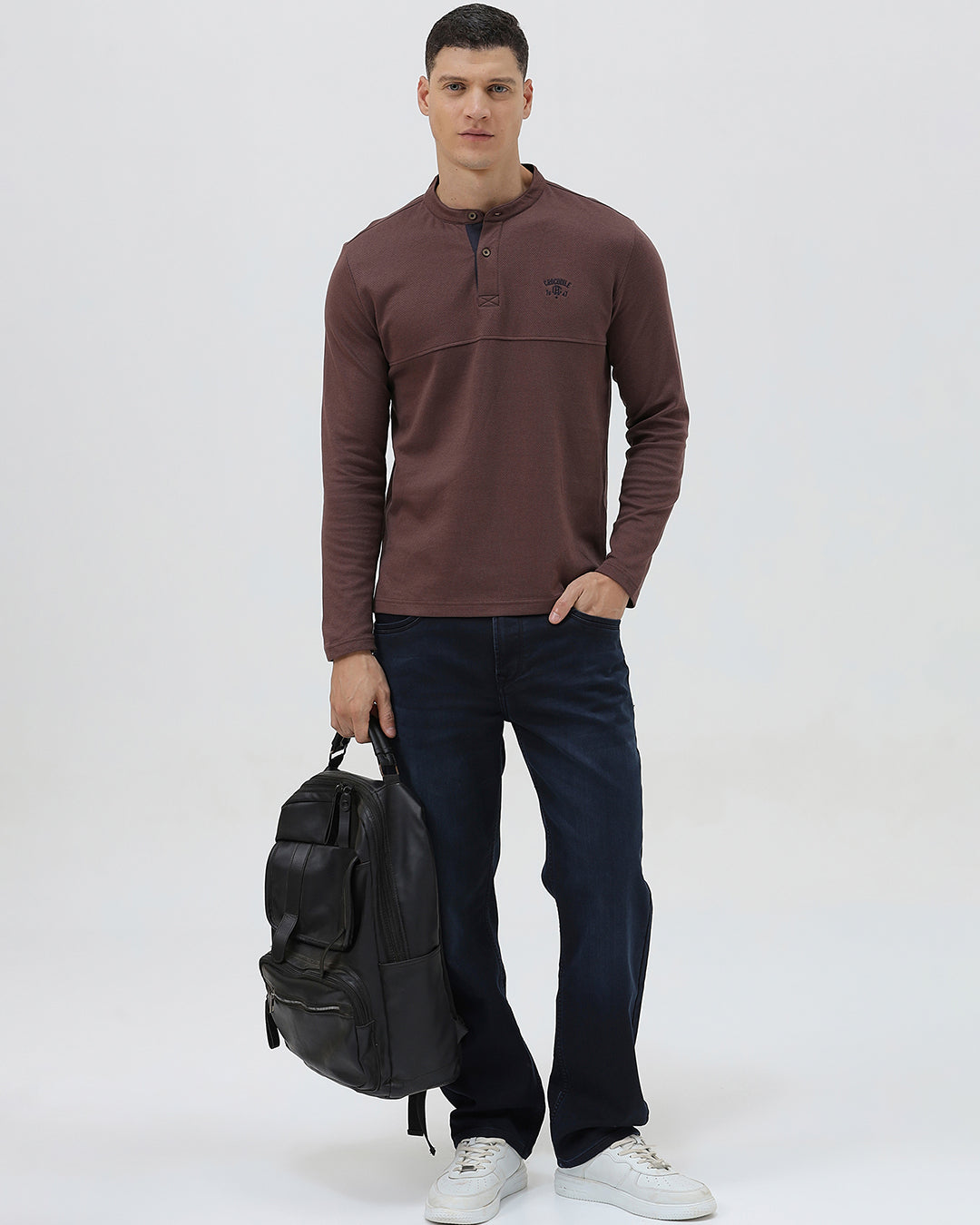 A man wearing a brown long-sleeve henley shirt with a small embroidered logo on the left chest, paired with dark blue jeans, standing against a light gray background.