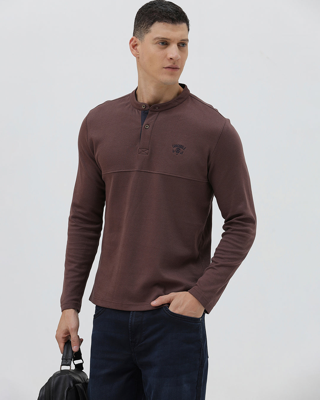 A man wearing a brown long-sleeve henley shirt with a small embroidered logo on the left chest, paired with dark blue jeans, standing against a light gray background.