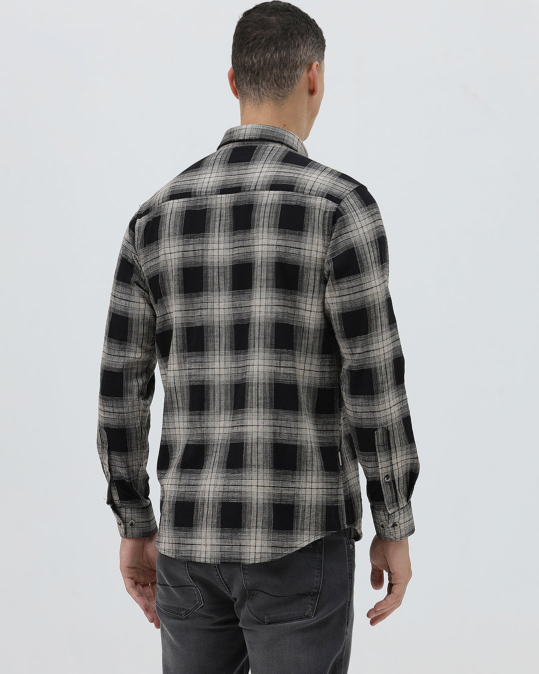 A man wearing a long-sleeve button-up plaid shirt in black, gray, and white, paired with dark gray jeans, standing against a light gray studio background.