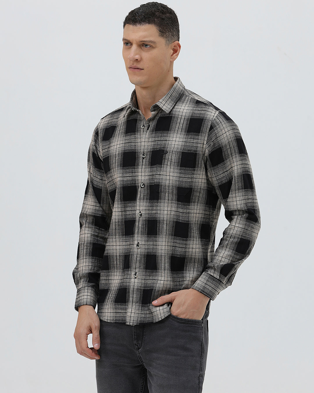 A man wearing a long-sleeve button-up plaid shirt in black, gray, and white, paired with dark gray jeans, standing against a light gray studio background.