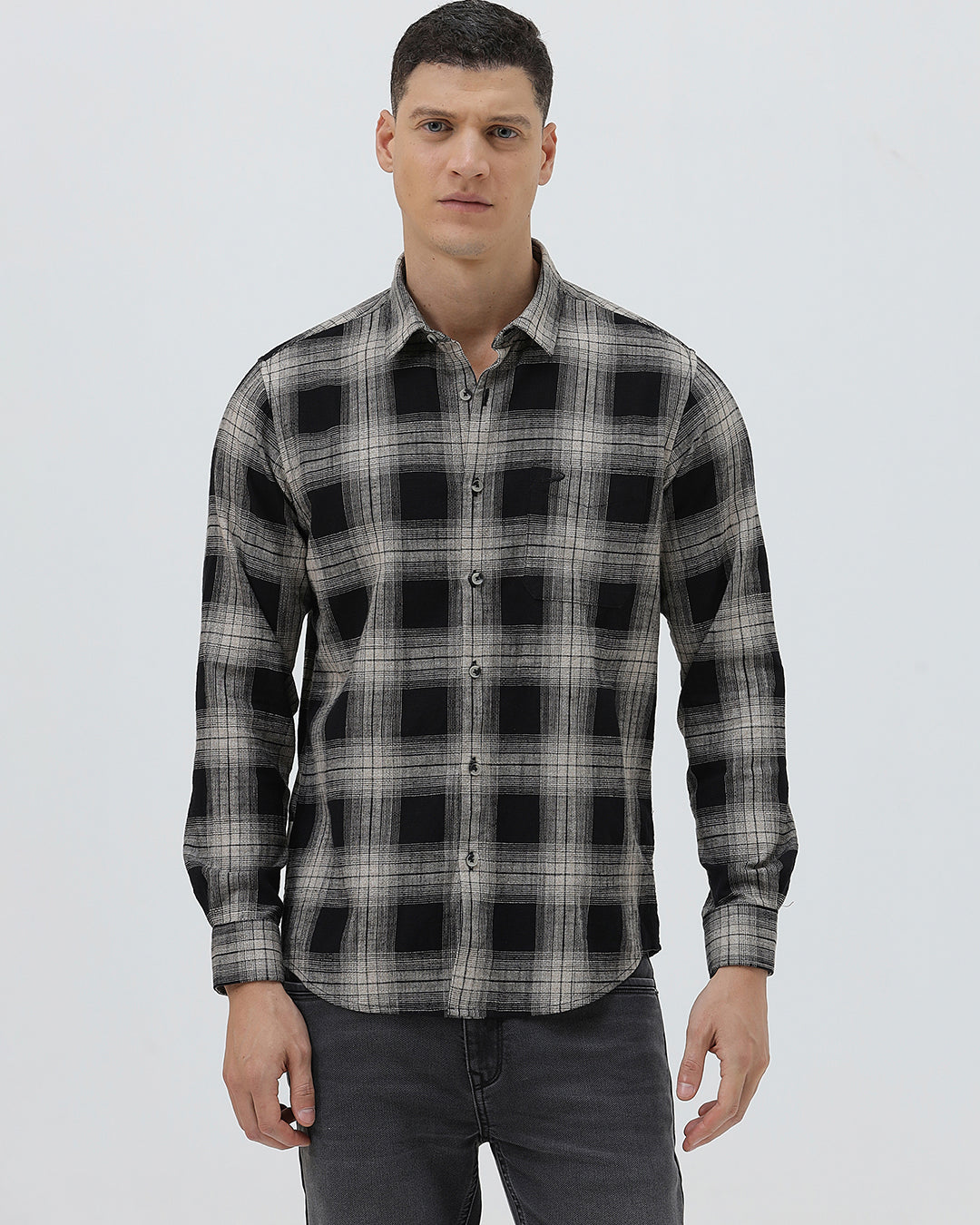A man wearing a long-sleeve button-up plaid shirt in black, gray, and white, paired with dark gray jeans, standing against a light gray studio background.