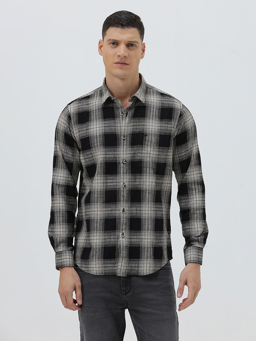 A man wearing a long-sleeve button-up plaid shirt in black, gray, and white, paired with dark gray jeans, standing against a light gray studio background.