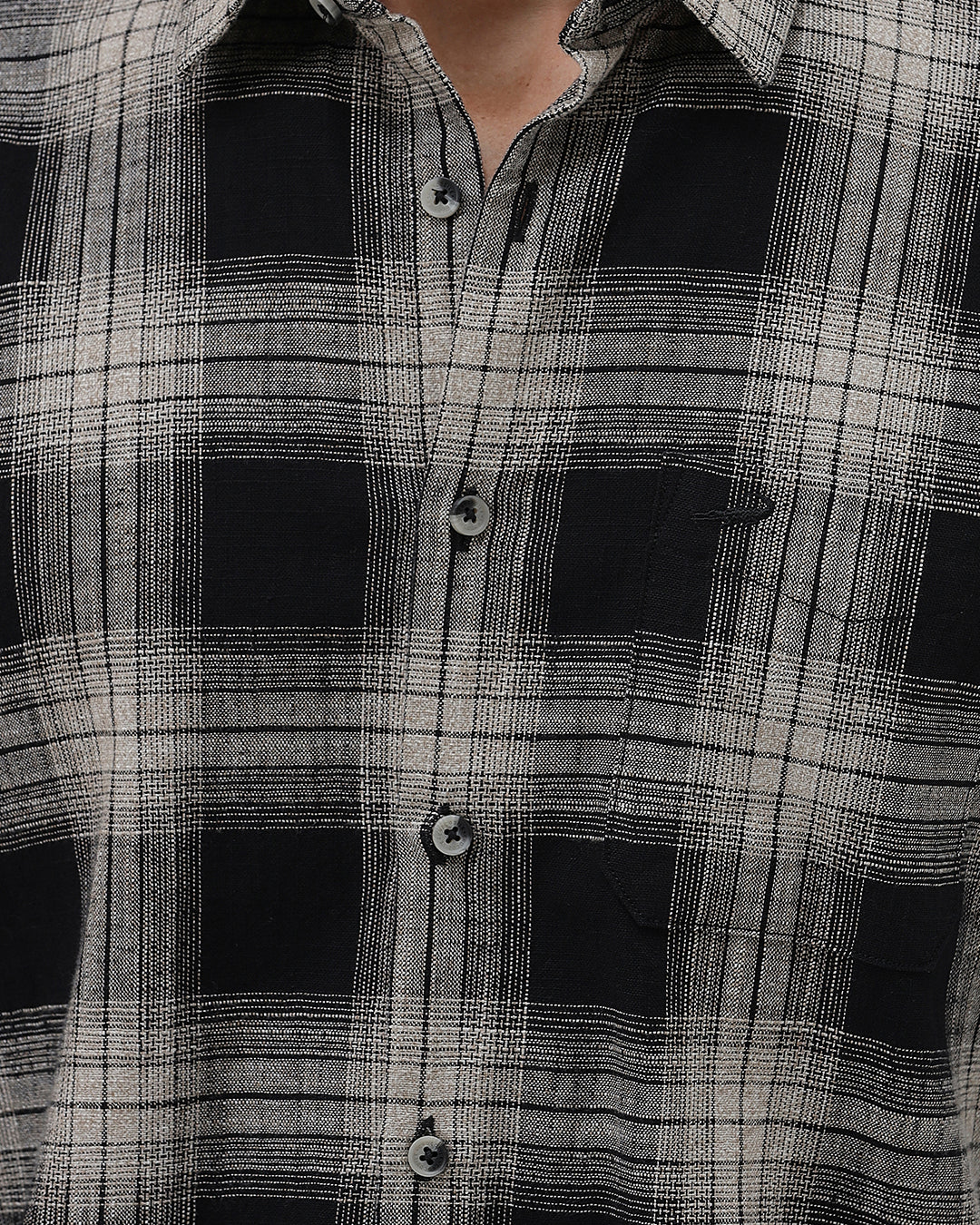 A man wearing a long-sleeve button-up plaid shirt in black, gray, and white, paired with dark gray jeans, standing against a light gray studio background.