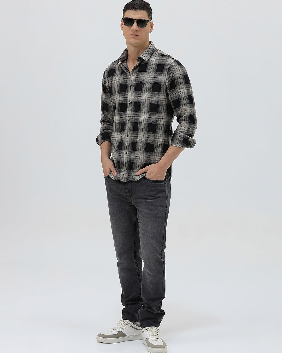 A man wearing a long-sleeve button-up plaid shirt in black, gray, and white, paired with dark gray jeans, standing against a light gray studio background.