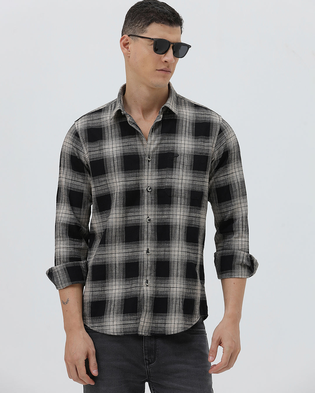 A man wearing a long-sleeve button-up plaid shirt in black, gray, and white, paired with dark gray jeans, standing against a light gray studio background.