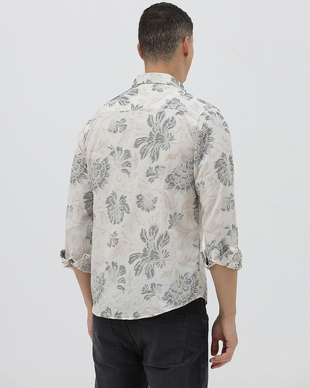 A man wearing a long-sleeve button-up shirt with a gray floral pattern on a cream-colored base, paired with dark gray trousers, against a plain light gray background.