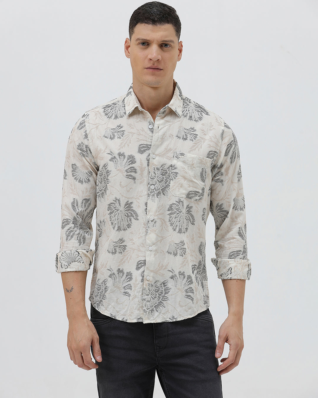A man wearing a long-sleeve button-up shirt with a gray floral pattern on a cream-colored base, paired with dark gray trousers, against a plain light gray background.