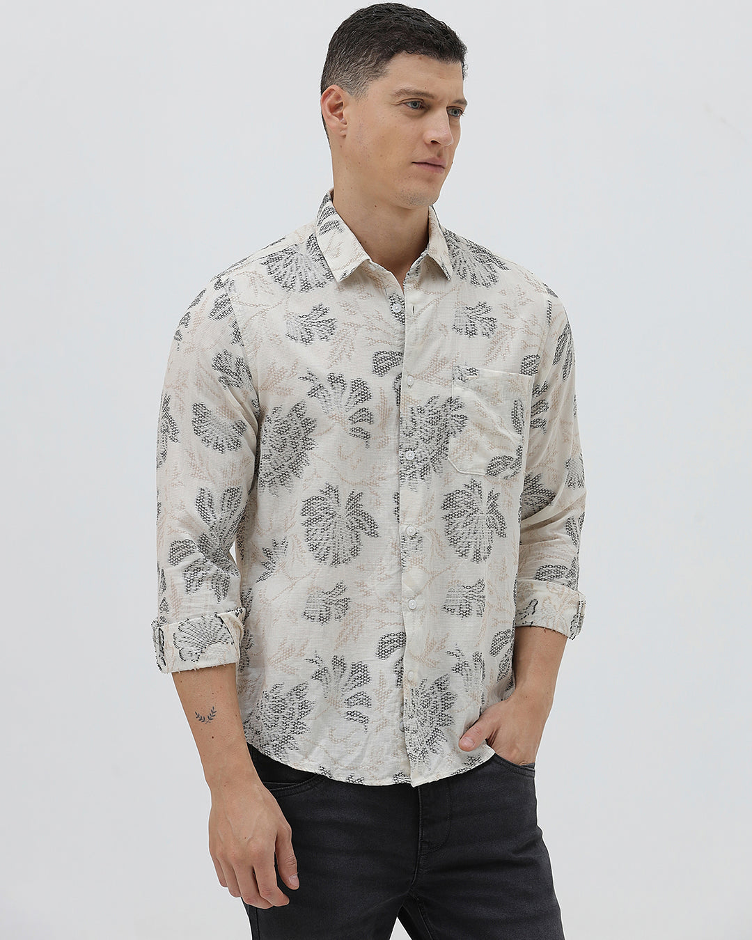 A man wearing a long-sleeve button-up shirt with a gray floral pattern on a cream-colored base, paired with dark gray trousers, against a plain light gray background.