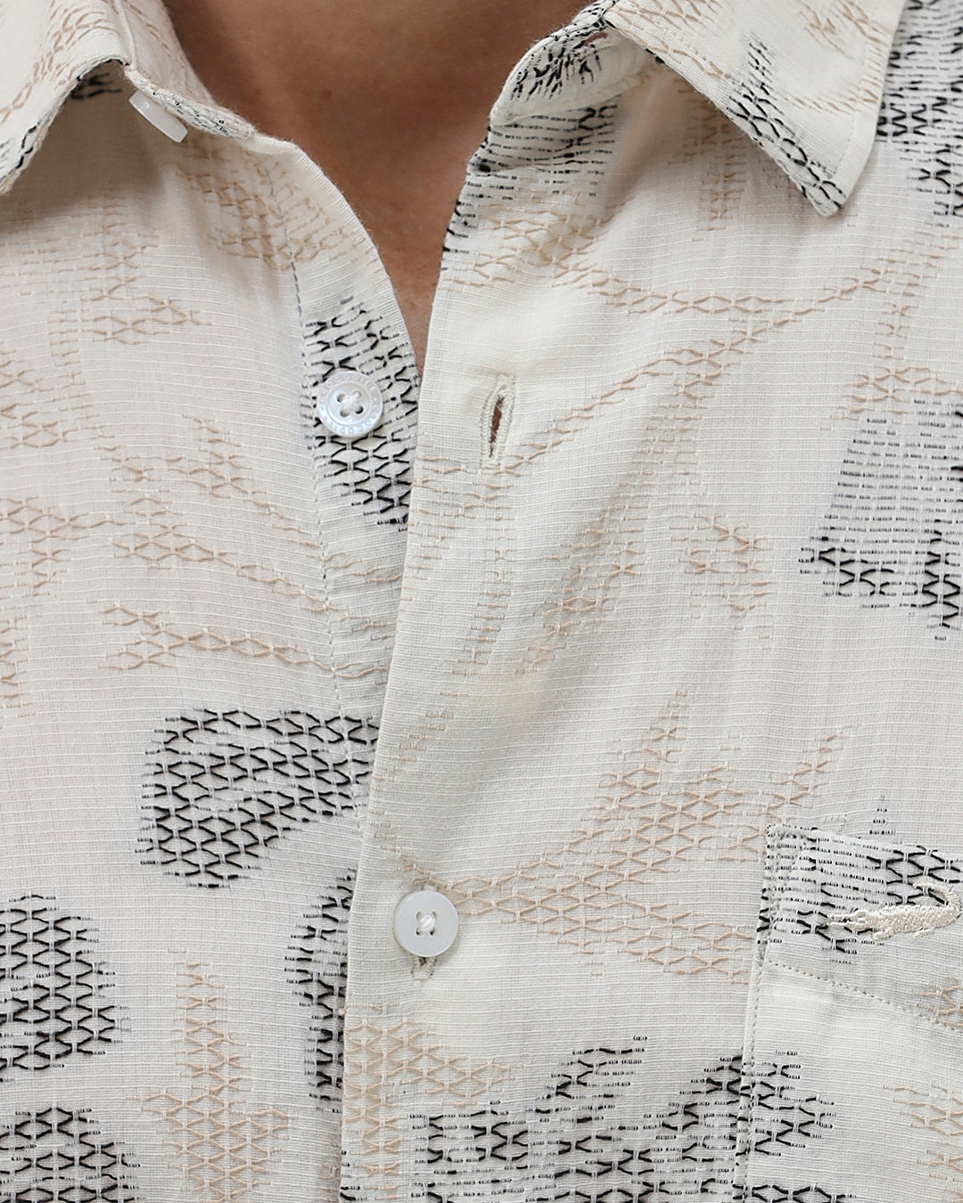 A man wearing a long-sleeve button-up shirt with a gray floral pattern on a cream-colored base, paired with dark gray trousers, against a plain light gray background.
