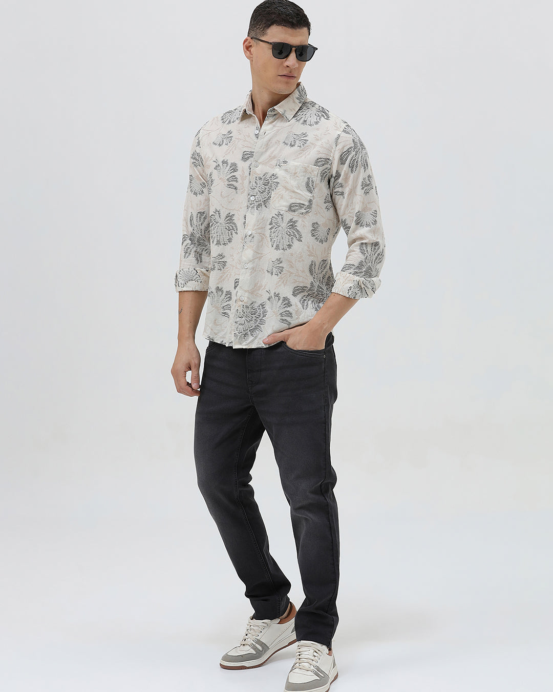 A man wearing a long-sleeve button-up shirt with a gray floral pattern on a cream-colored base, paired with dark gray trousers, against a plain light gray background.
