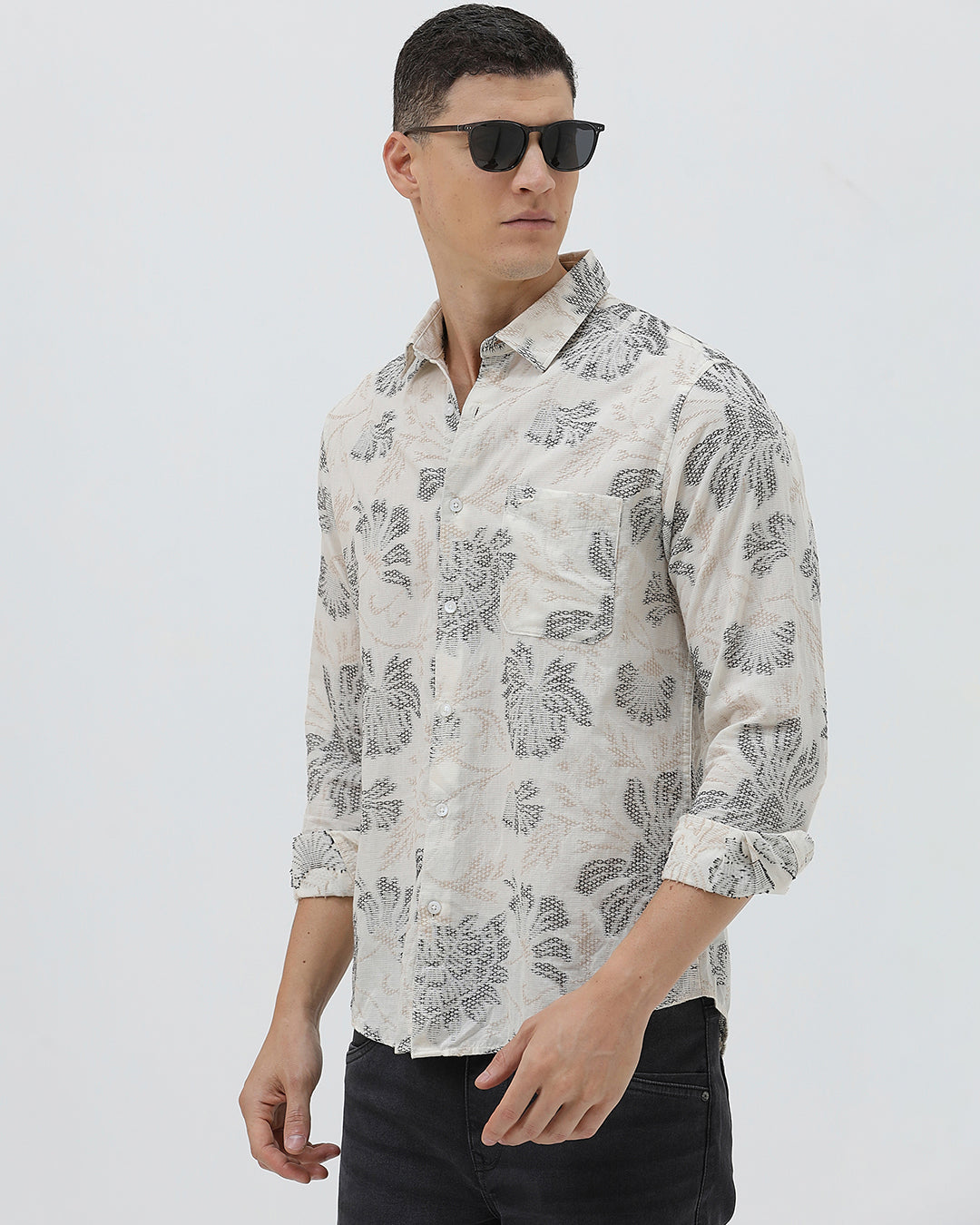 A man wearing a long-sleeve button-up shirt with a gray floral pattern on a cream-colored base, paired with dark gray trousers, against a plain light gray background.