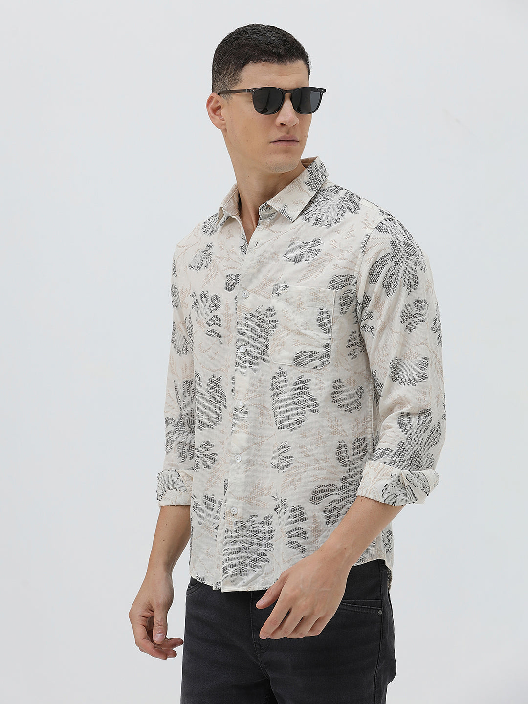 A man wearing a long-sleeve button-up shirt with a gray floral pattern on a cream-colored base, paired with dark gray trousers, against a plain light gray background.