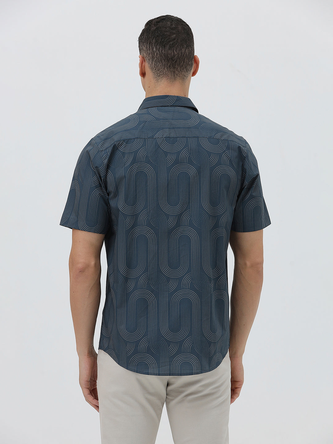 A man wearing a short-sleeve button-up shirt with a dark blue and gray abstract wavy pattern, paired with light beige trousers, standing against a plain light gray background.