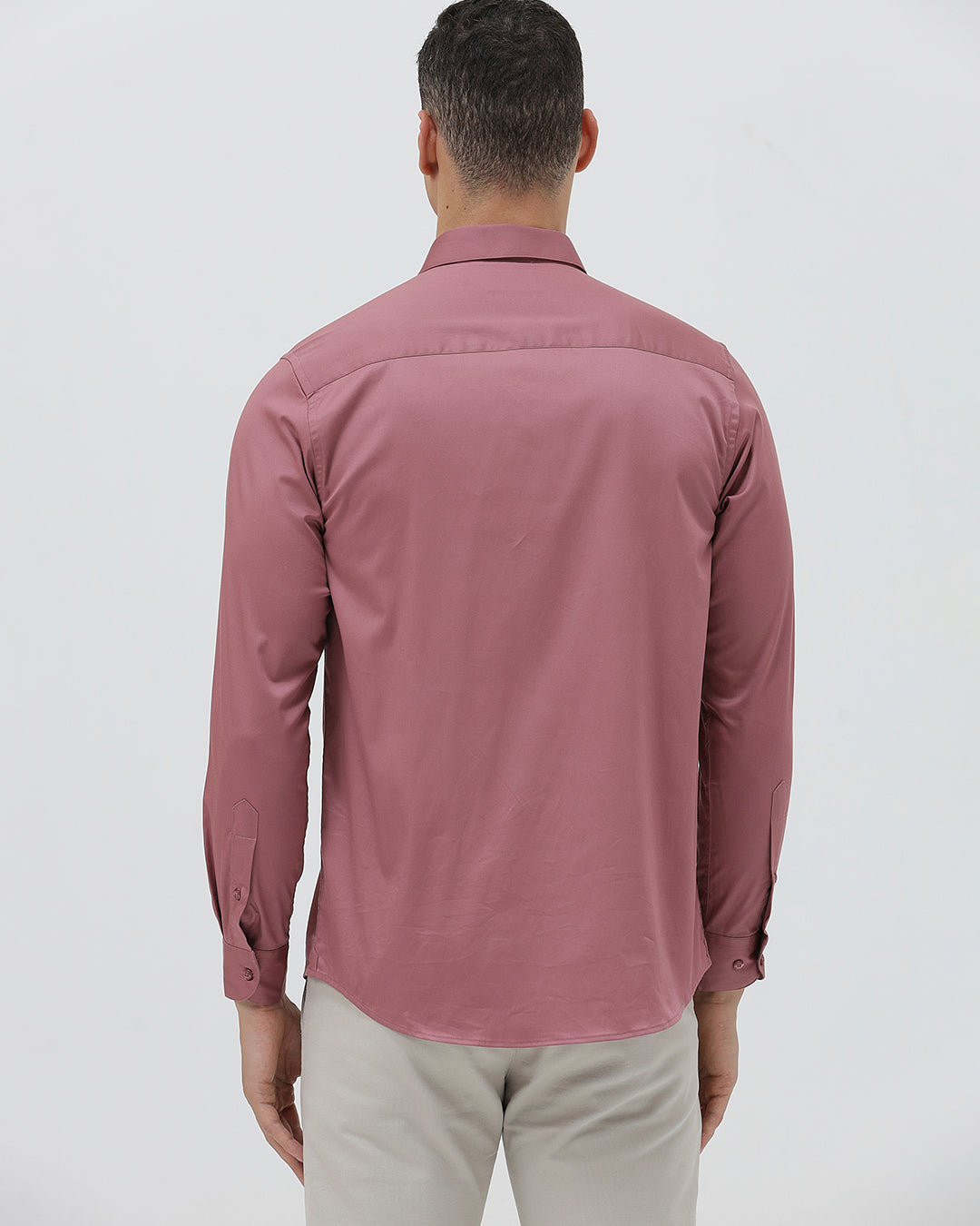 A man wearing a dusty rose long-sleeve button-down shirt and light beige trousers, standing against a light gray studio background.
