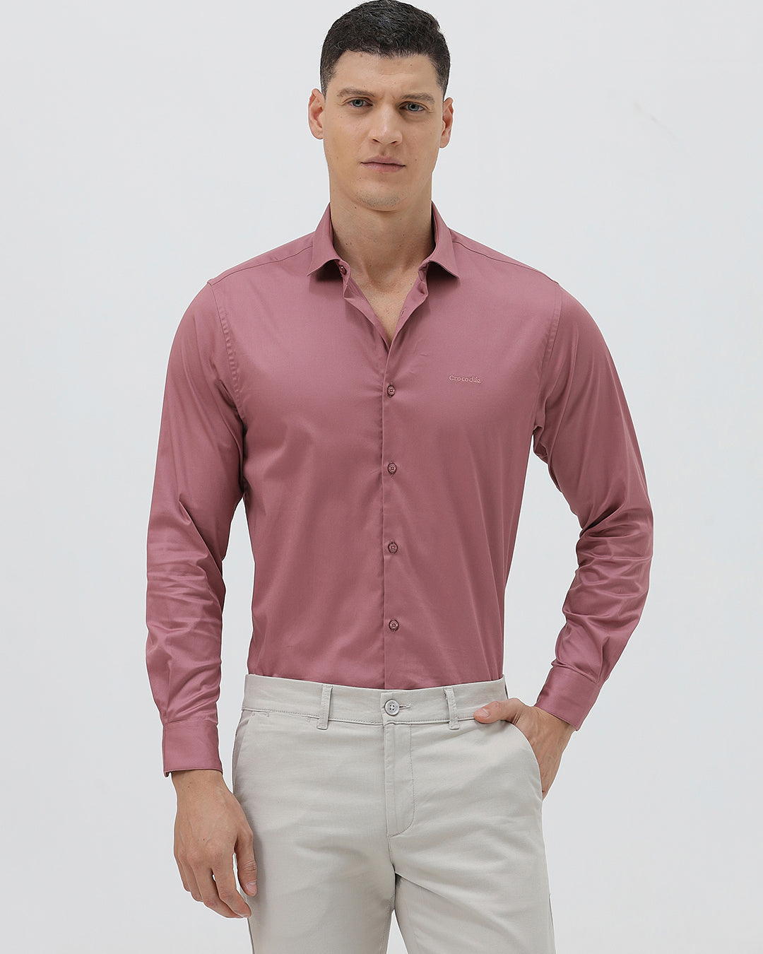 A man wearing a dusty rose long-sleeve button-down shirt and light beige trousers, standing against a light gray studio background.