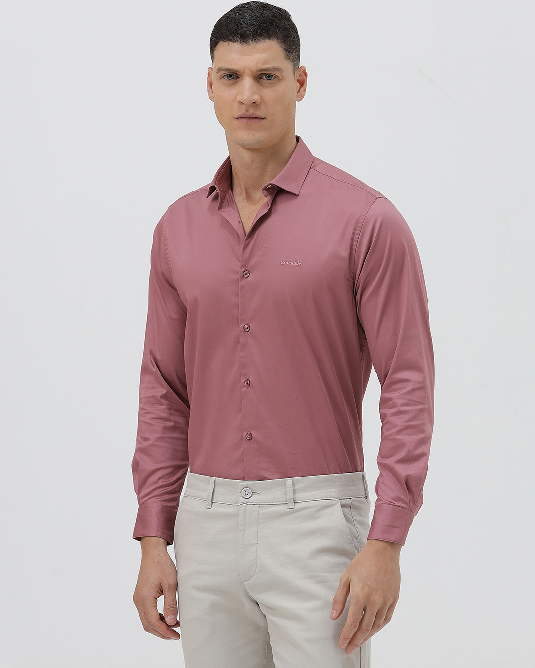 A man wearing a dusty rose long-sleeve button-down shirt and light beige trousers, standing against a light gray studio background.