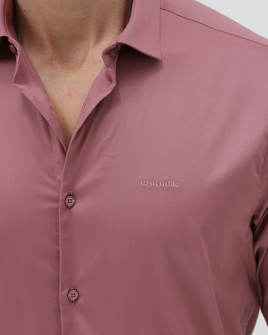 A man wearing a dusty rose long-sleeve button-down shirt and light beige trousers, standing against a light gray studio background.