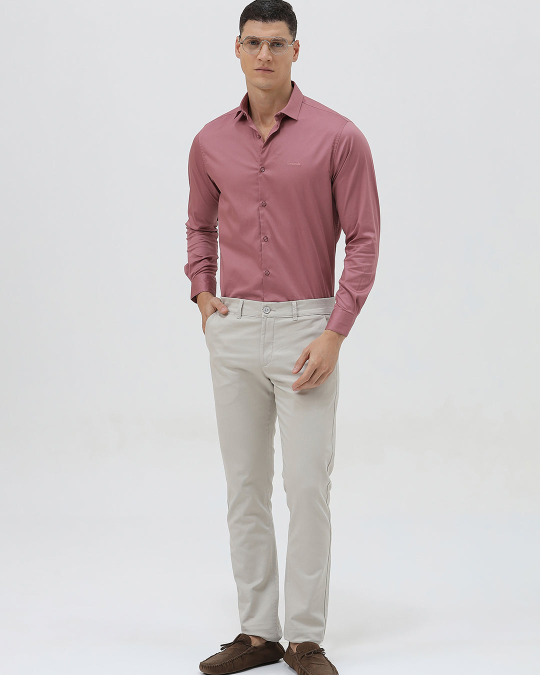 A man wearing a dusty rose long-sleeve button-down shirt and light beige trousers, standing against a light gray studio background.