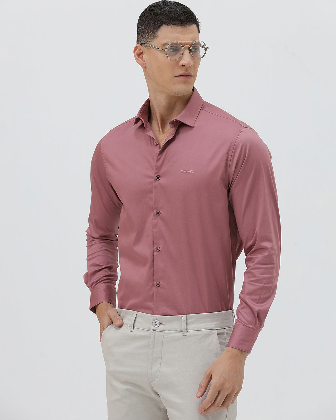 A man wearing a dusty rose long-sleeve button-down shirt and light beige trousers, standing against a light gray studio background.