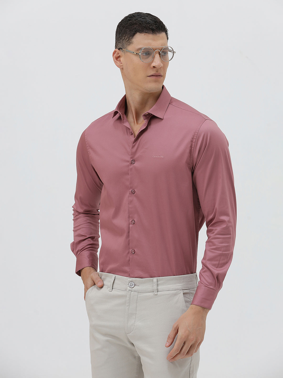 A man wearing a dusty rose long-sleeve button-down shirt and light beige trousers, standing against a light gray studio background.