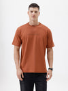 A man wearing a rust-orange crewneck t-shirt with a subtle tonal 'ELECTRIC' logo across the chest, paired with black pants, photographed against a clean white studio background.