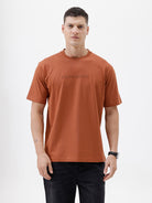 A man wearing a rust-orange crewneck t-shirt with a subtle tonal 'ELECTRIC' logo across the chest, paired with black pants, photographed against a clean white studio background.