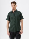 A man wearing a dark forest green short-sleeve button-up shirt with a chest pocket, paired with black jeans, standing against a plain white studio background.