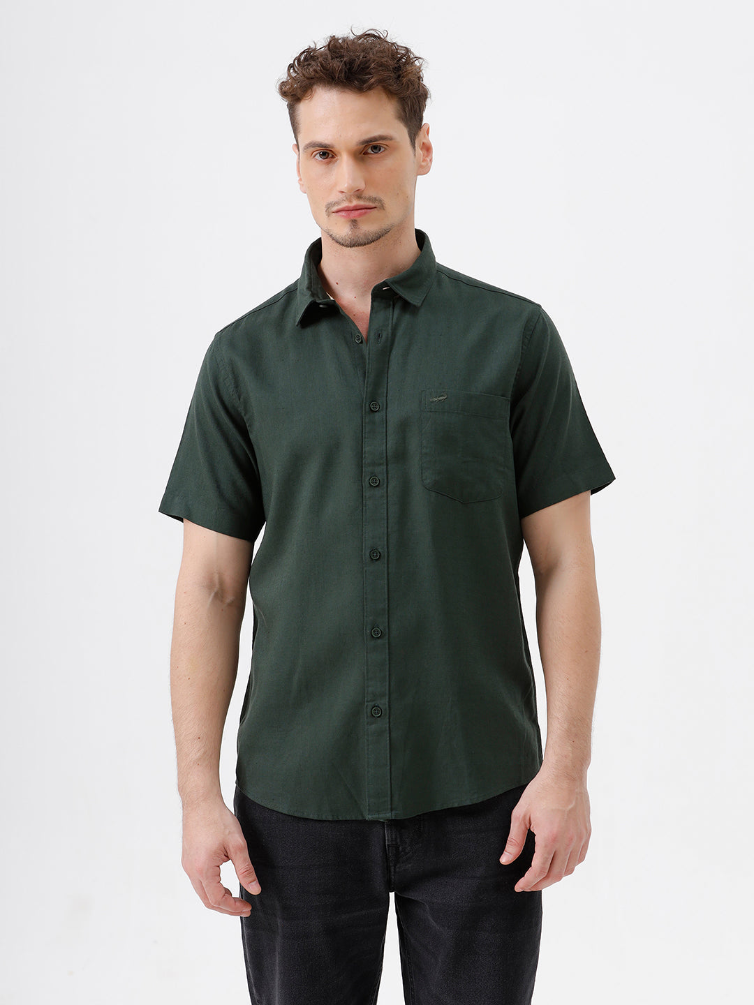 A man wearing a dark forest green short-sleeve button-up shirt with a chest pocket, paired with black jeans, standing against a plain white studio background.