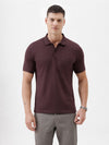 A man wearing a short-sleeve polo shirt in dark burgundy with a chest pocket, paired with gray trousers, standing against a plain light gray background.