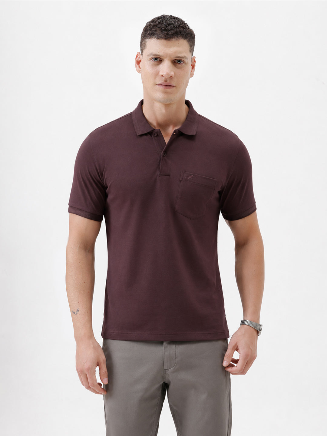 A man wearing a short-sleeve polo shirt in dark burgundy with a chest pocket, paired with gray trousers, standing against a plain light gray background.