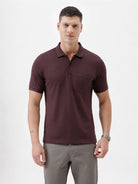 A man wearing a short-sleeve polo shirt in dark burgundy with a chest pocket, paired with gray trousers, standing against a plain light gray background.