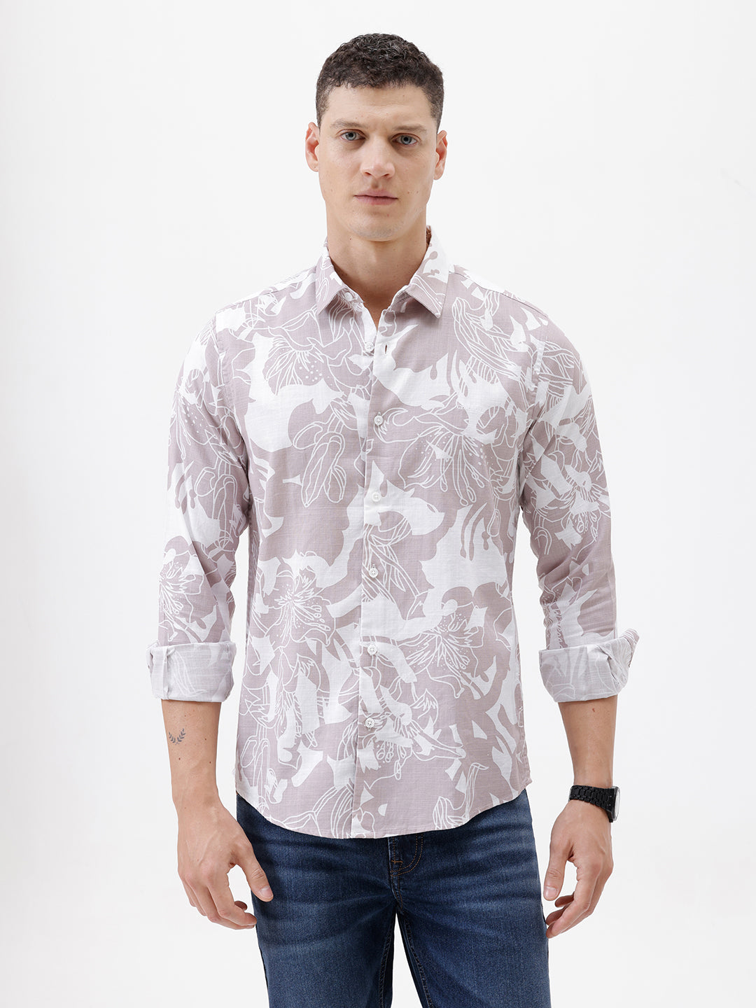 A man wearing a long-sleeve button-up shirt with a light gray and white abstract floral pattern, paired with dark blue jeans, standing against a plain white background.
