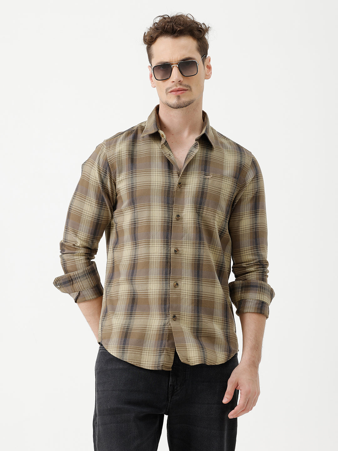 A man wearing a brown and beige plaid long-sleeve button-down shirt with dark sunglasses and black jeans, standing against a plain white studio background.