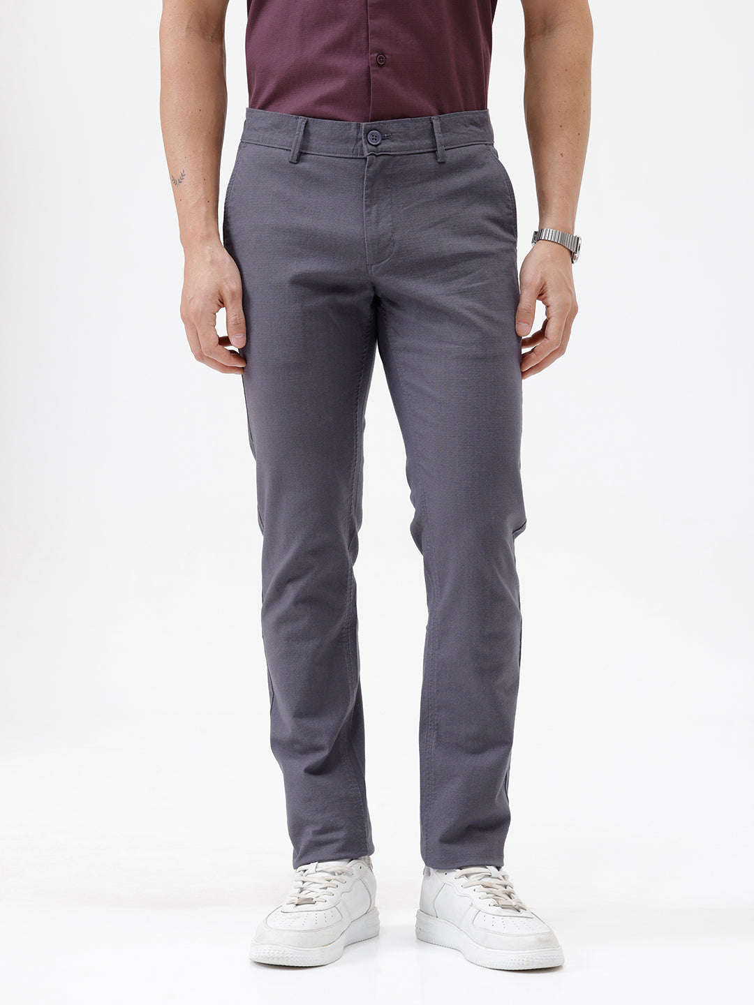 A person wearing slim-fit charcoal gray chino pants, paired with a dark purple shirt and white sneakers, standing against a plain white studio background.
