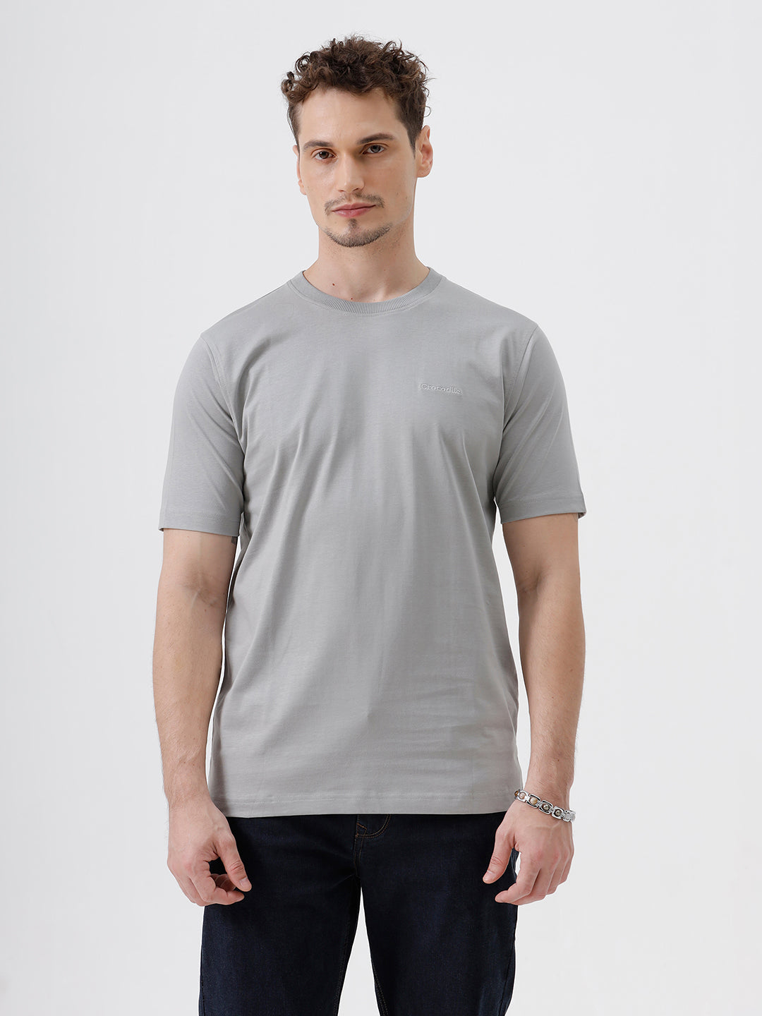 A man wearing a light gray short-sleeve crewneck t-shirt with a subtle tonal chest logo, paired with dark jeans, standing against a light gray studio background.