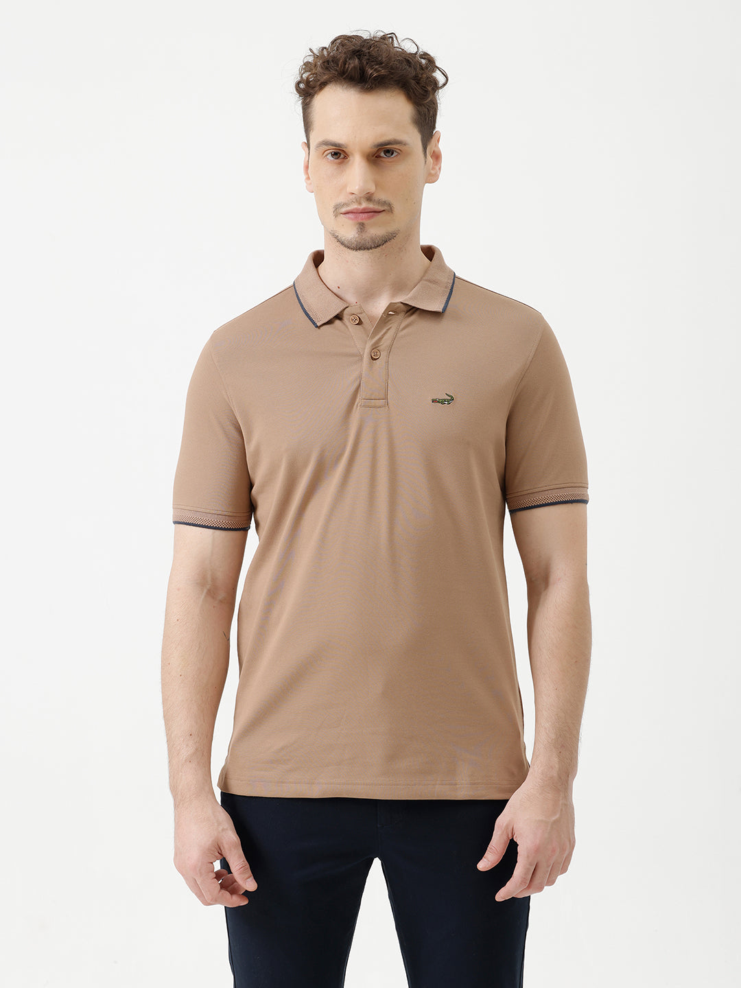 A man wearing a tan short-sleeve polo shirt with dark blue piping on the collar and sleeve cuffs, paired with dark navy pants, against a plain light gray background.