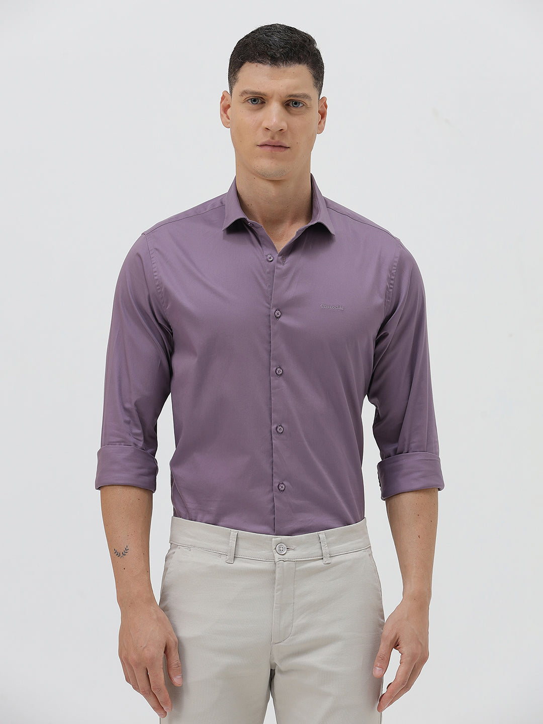 A man wearing a purple long-sleeve button-down shirt with rolled-up sleeves and light beige trousers, standing against a plain light gray background.