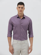 A man wearing a purple long-sleeve button-down shirt with rolled-up sleeves and light beige trousers, standing against a plain light gray background.