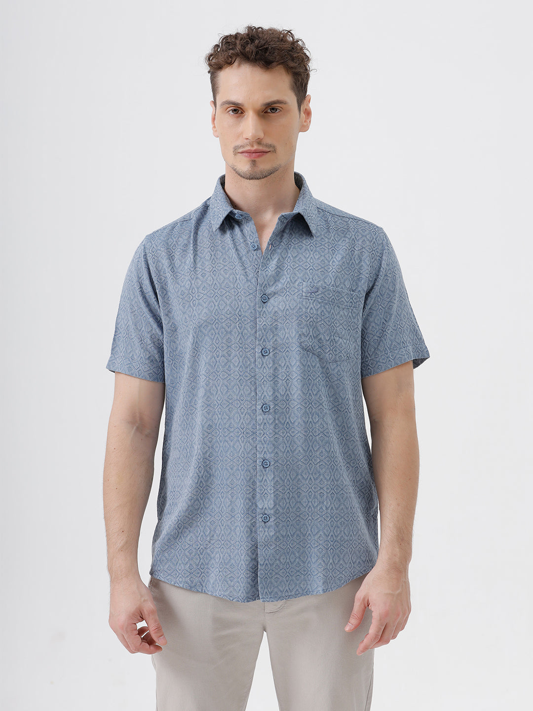 A man wearing a short-sleeve button-up shirt with a fine blue and white geometric pattern, paired with light beige trousers, standing against a plain light gray background.