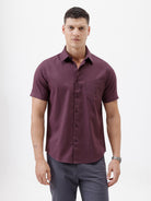 A man modeling a short-sleeve button-up shirt in a deep plum purple color, paired with gray trousers, against a plain white studio background.